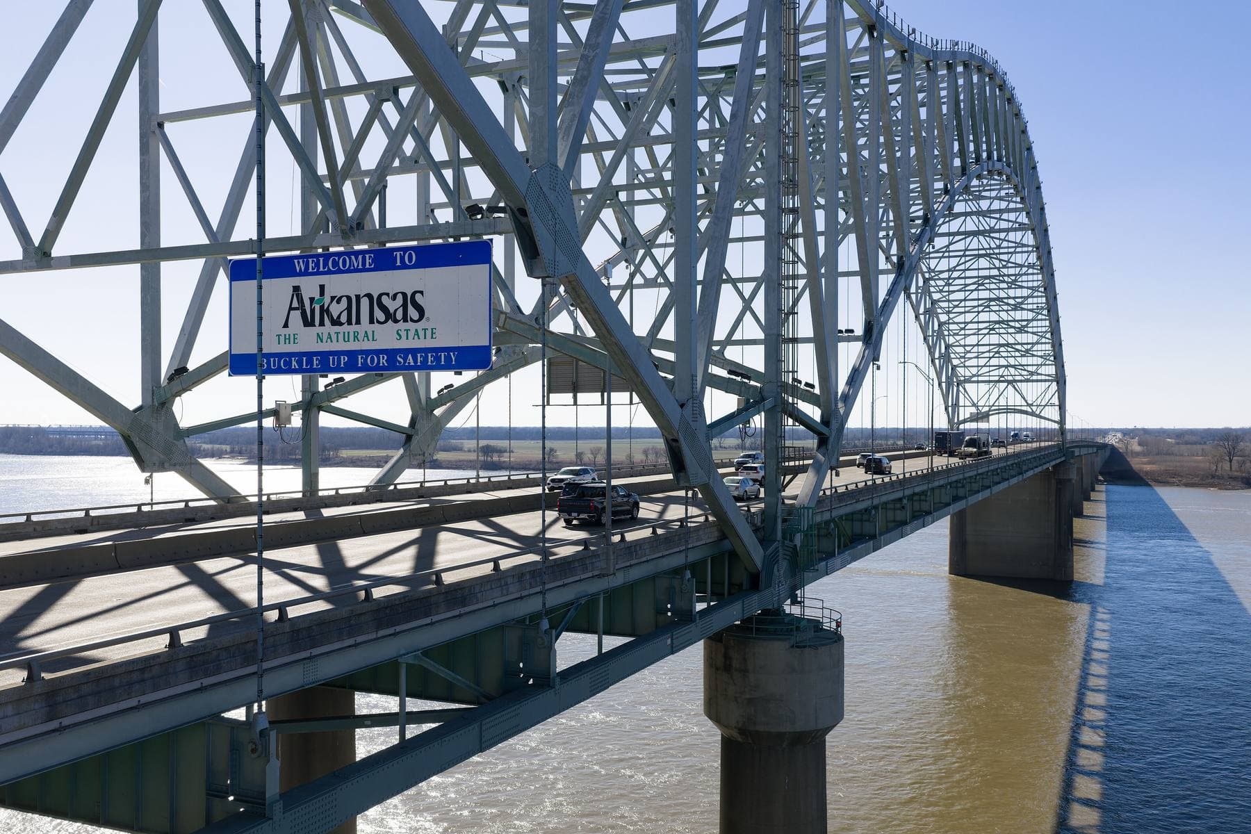 A blue-bordered 'Welcome to Arkansas, The Natural State' sign mounted on the steel arch of the Interstate 40 bridge over the Mississippi River