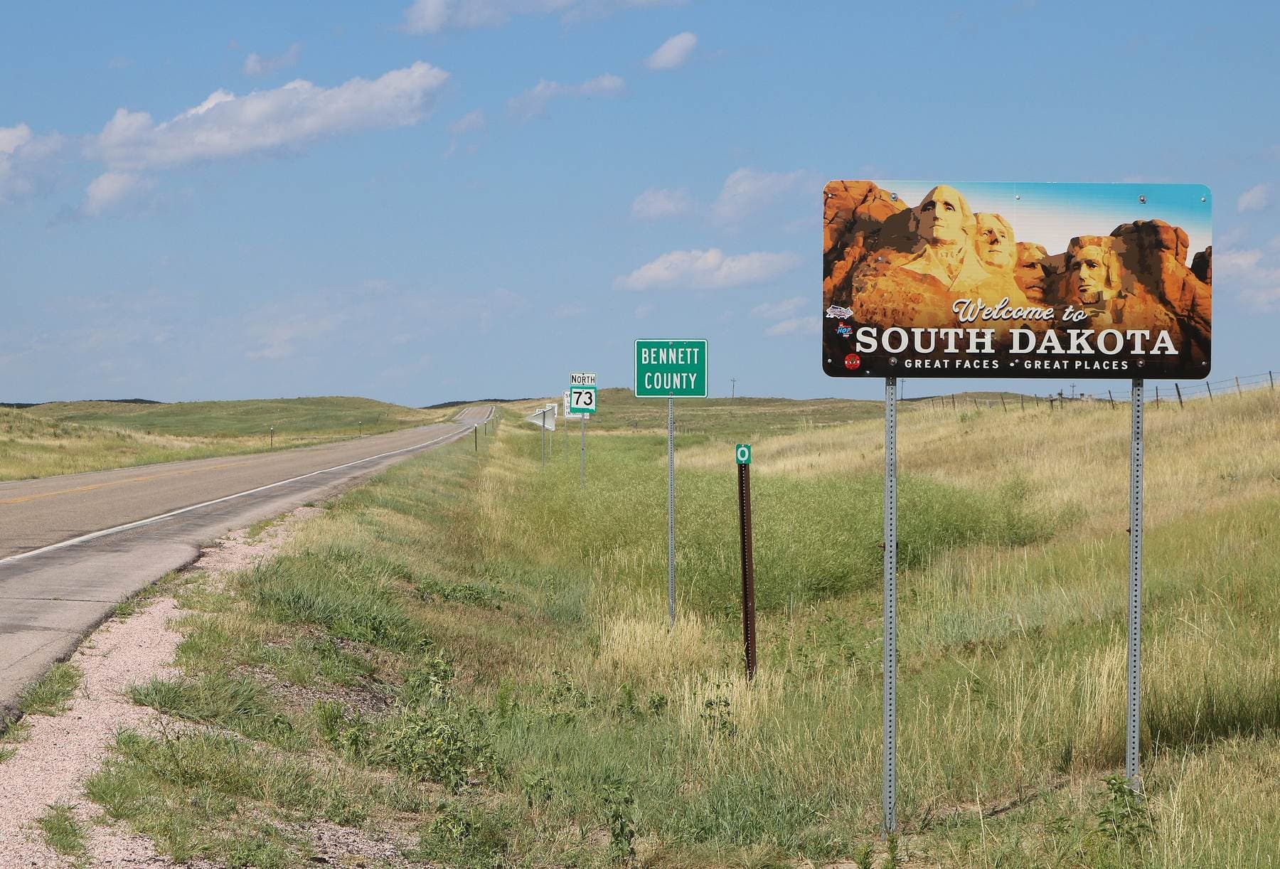 A 'Welcome to South Dakota' sign beside a two-lane highway at the Nebraska state line, with open grassland and a blue sky